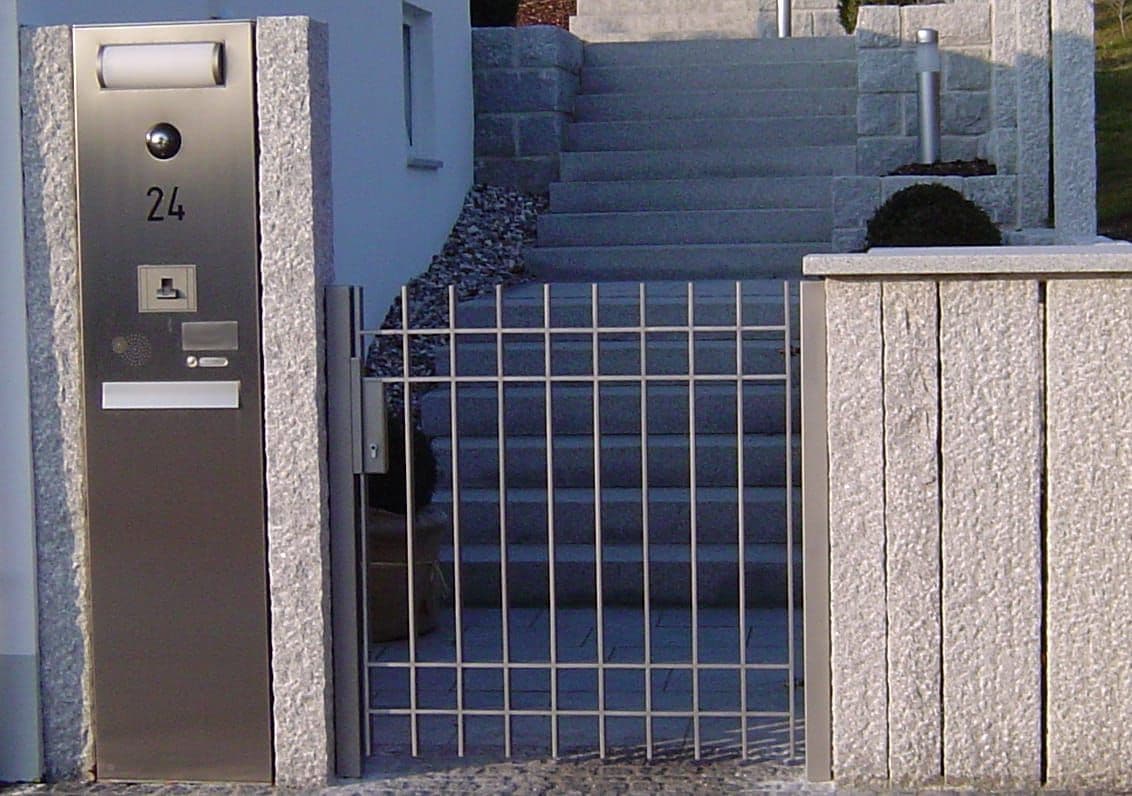 Briefkasten- und Klingelsäule aus Edelstahl neben einem Metalltor